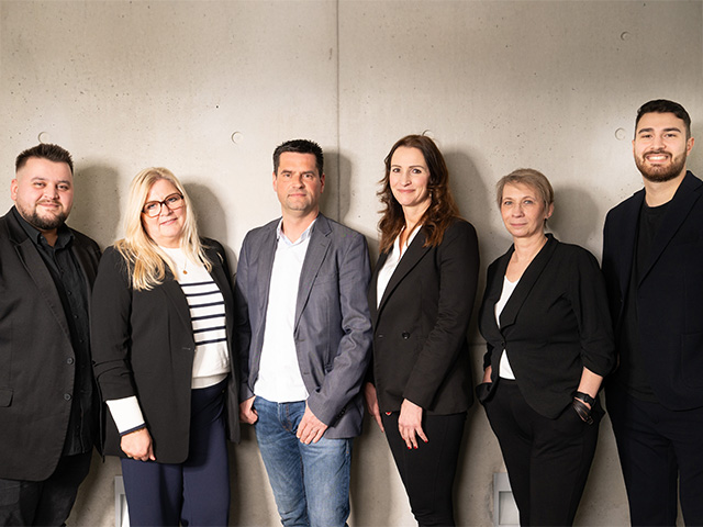 Sechs Personen stehen nebeneinander vor einer hellen Betonwand. Alle tragen formelle Kleidung und blicken freundlich in die Kamera. Die Aufnahme vermittelt ein professionelles, einladendes Team.