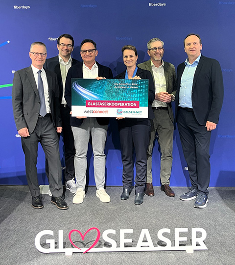 Sechs Personen in Businesskleidung stehen gemeinsam vor einer blauen Wand mit dem Schriftzug 'fiberdays'. Zwei von ihnen halten ein Schild mit der Aufschrift 'Glasfaserkooperation - Westconnect & Gelsen-Net'. Am unteren Bildrand ist der Schriftzug 'Gl♥seaser' zu sehen.